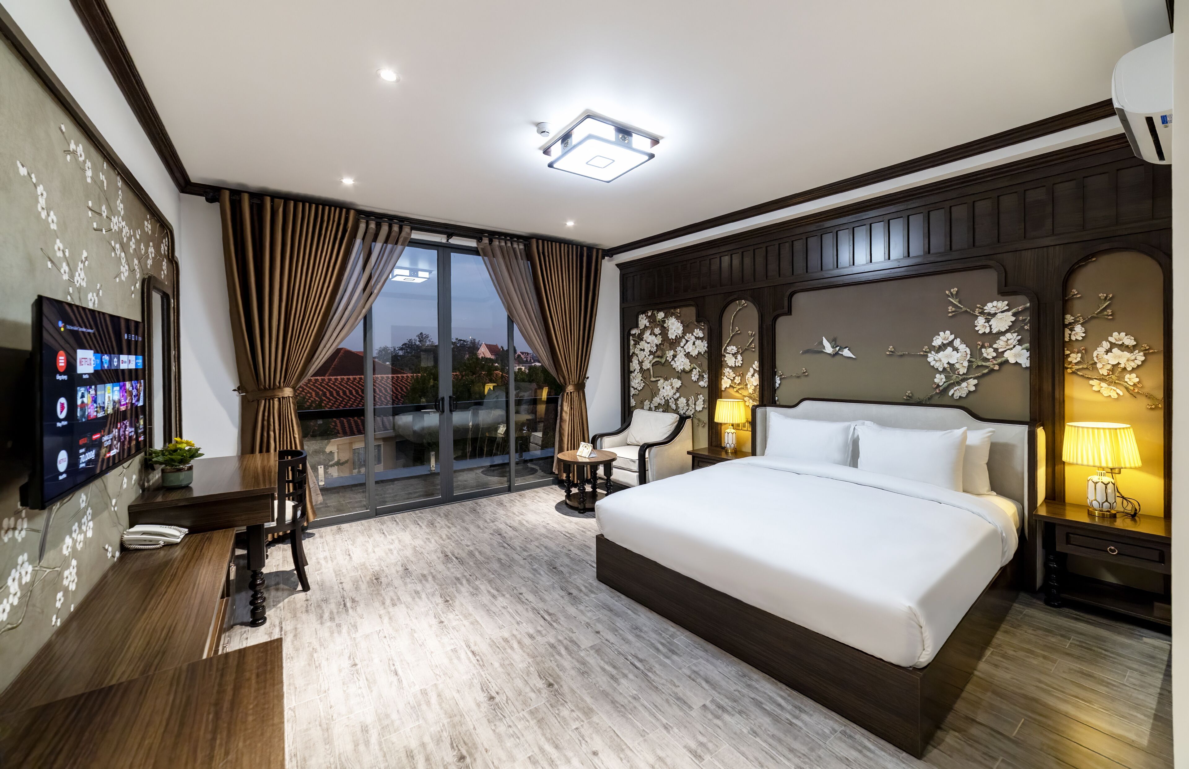 A modern hotel room with a large bed, a TV on the wall, a dining area, and a view of the outside.
