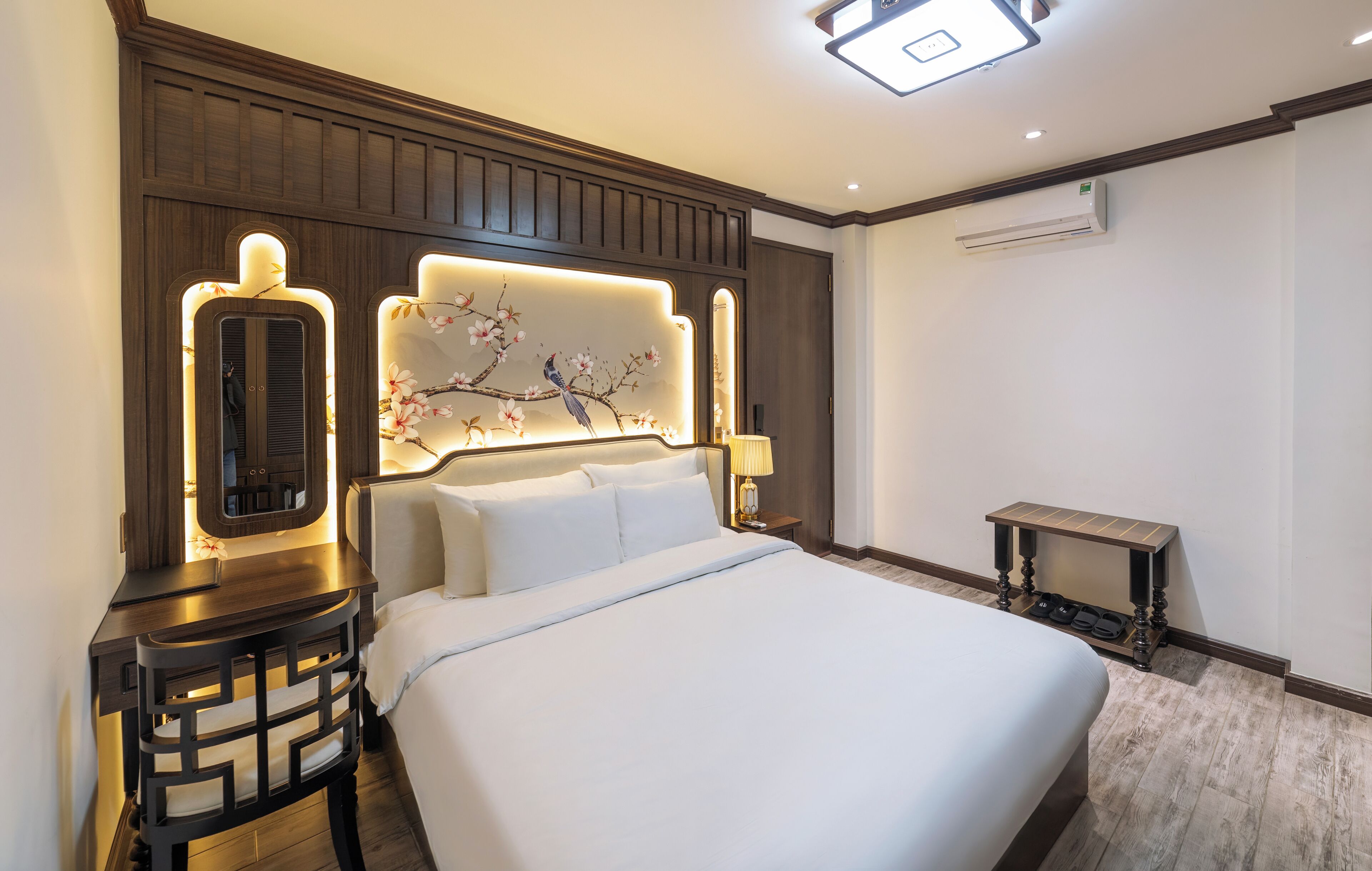 A hotel room with a large bed, a nightstand with a mirror, a wall-mounted air conditioner, and a decorative wall panel.