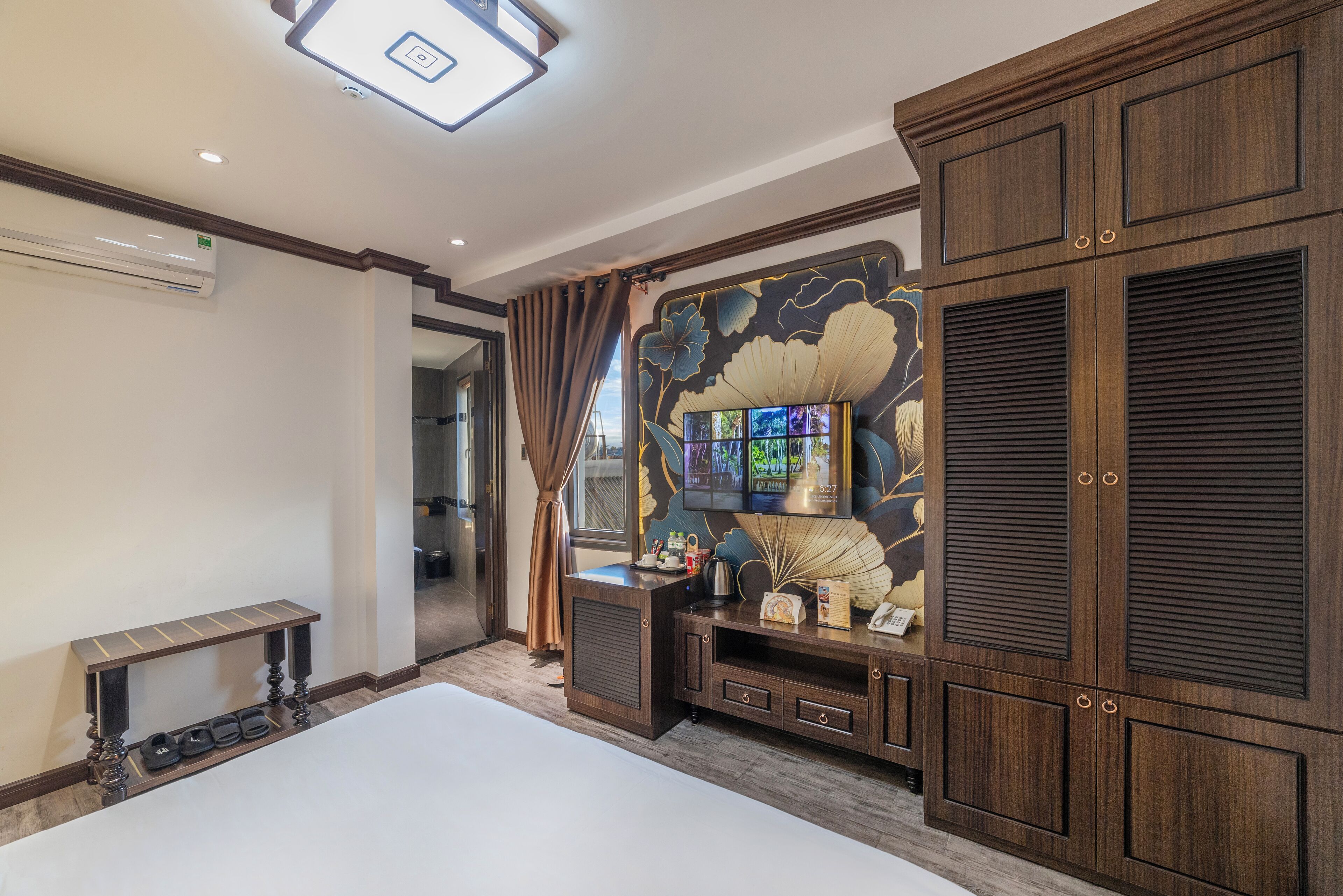 Une chambre d’hôtel avec un grand lit, un téléviseur fixé à un mur décoratif, des meubles en bois et une petite table avec des chaussons.