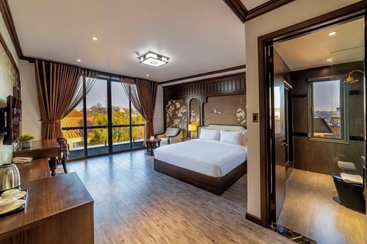 Een ruime slaapkamer met een groot bed, een bureau met stoel, en uitzicht op de stad door grote ramen.