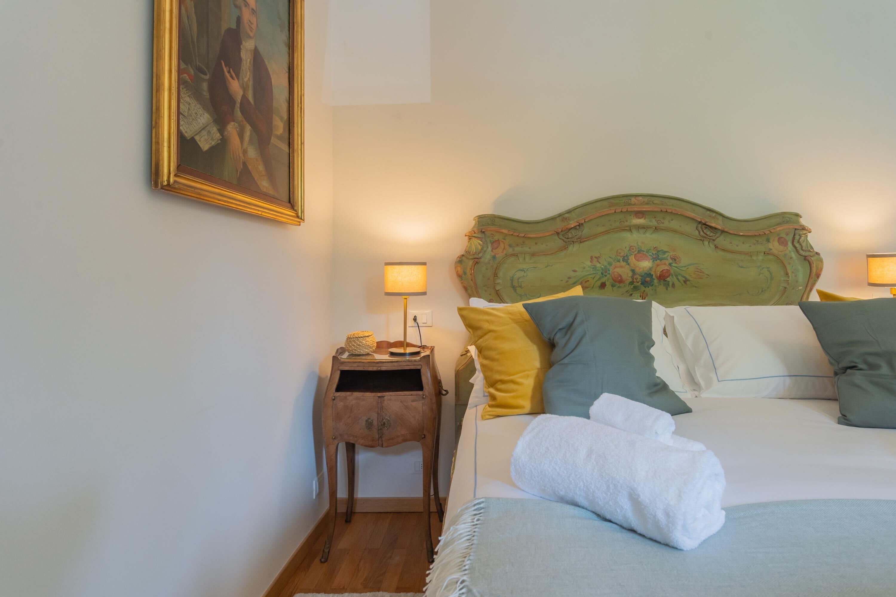 Uma cama bem arrumada com cabeceira verde, uma pequena mesa de cabeceira de madeira com abajur, e um quadro na parede.