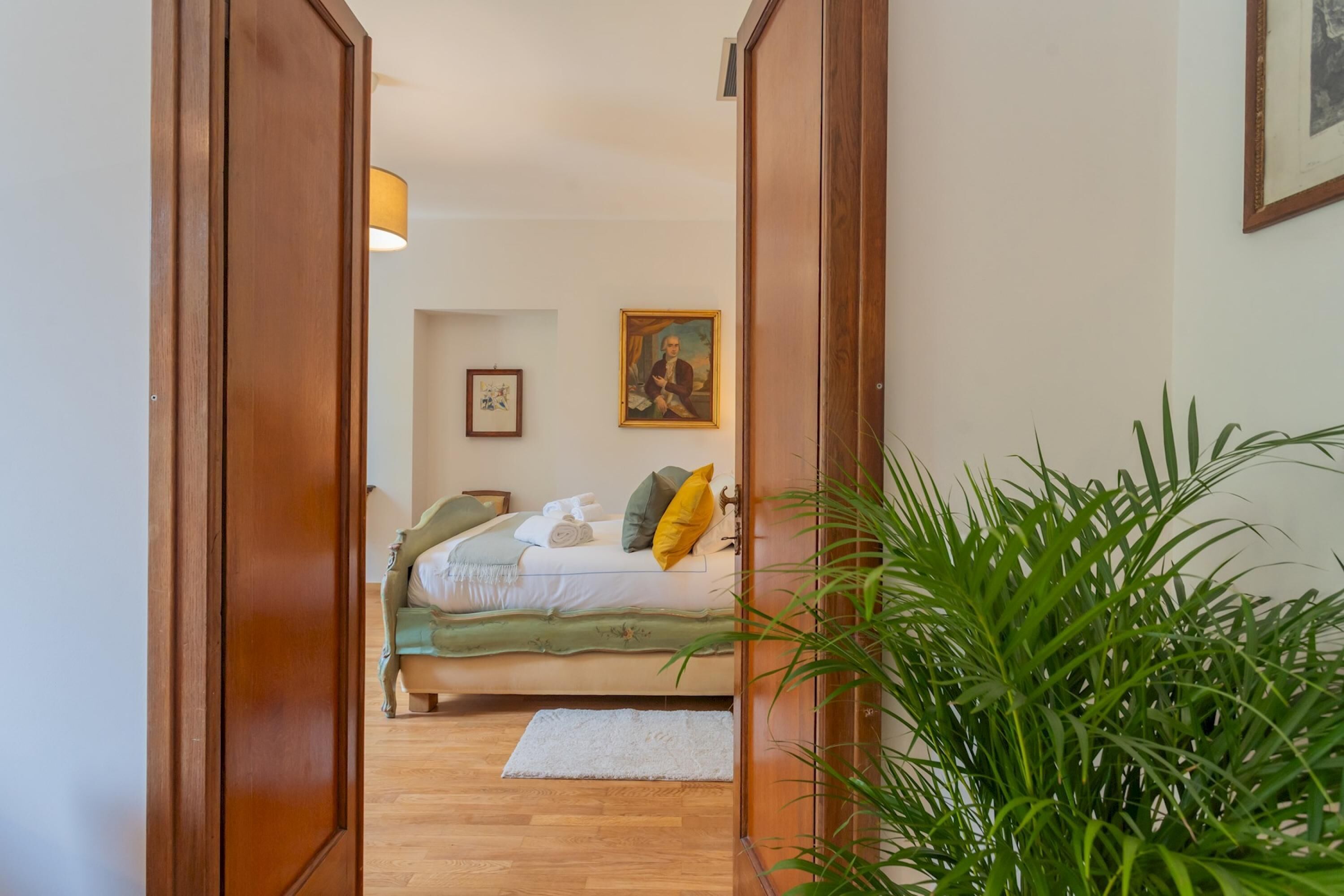 A bedroom with a bed, a bedside table, a lamp, a framed picture on the wall, and a potted plant.