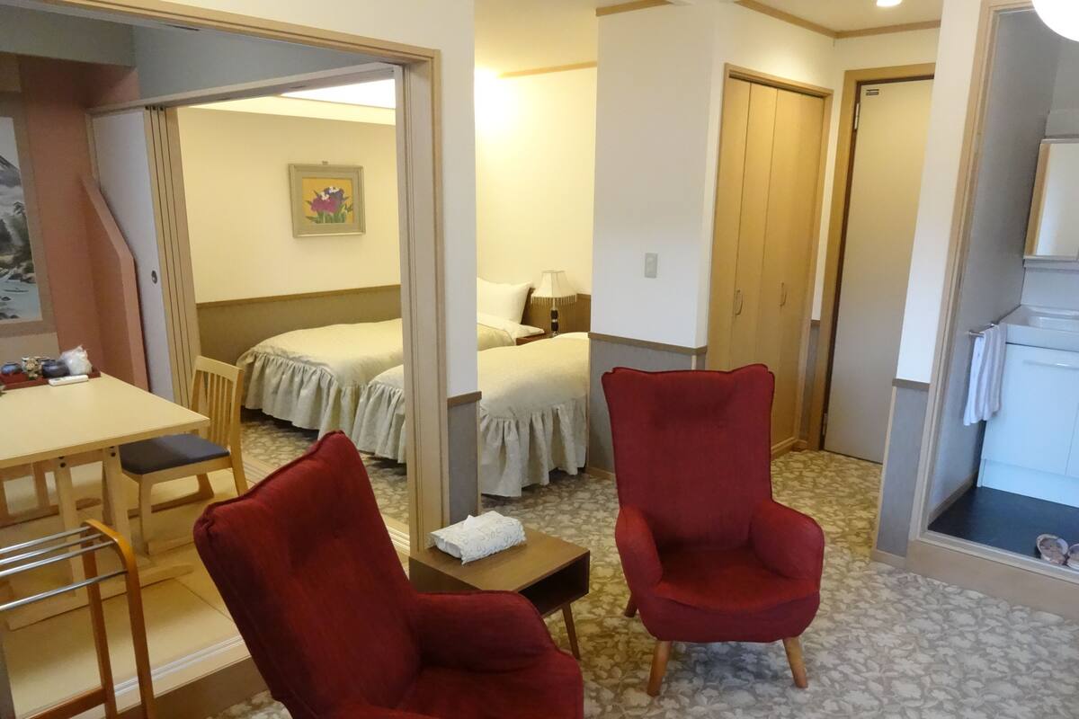 Quarto de hotel com cama, duas poltronas vermelhas, uma mesinha, uma escrivaninha e um banheiro.