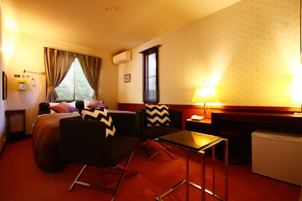 Quarto de hotel com cama, duas cadeiras, uma mesinha, escrivaninha, luminária, geladeira e janela com cortinas.
