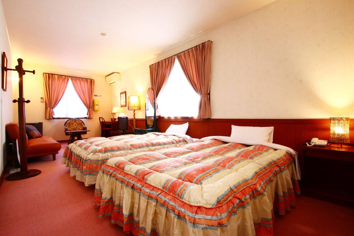Quarto de hotel com duas camas, uma escrivaninha, uma cadeira e uma mesa de cabeceira com abajur.