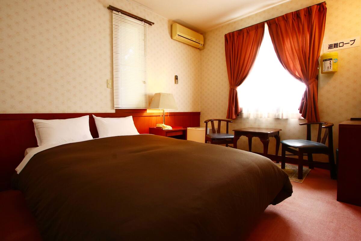 Quarto de hotel com uma cama grande, uma escrivaninha e uma cadeira. Há uma janela com cortinas, um ar condicionado e uma pequena mesa com uma luminária.