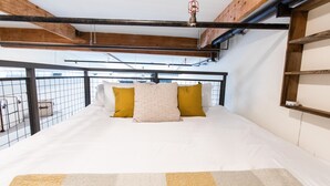Un lit avec une literie blanche et deux oreillers, un jaune et un blanc, dans une pièce dont les poutres en bois sont apparentes et qui est dotée d’une balustrade en métal.