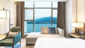 Habitación de hotel con una cama grande, un sofá, una silla y vistas a un cuerpo de agua y montañas.