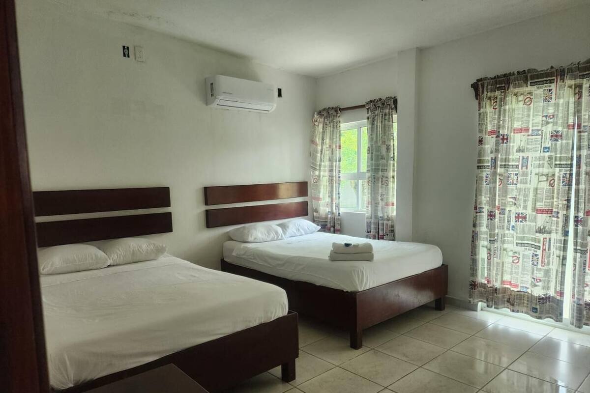A hotel room with two beds, a window with curtains, an air conditioning unit, and a tiled floor.