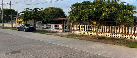 A street view with a parked car, a white fence, and a building with a gated entrance.