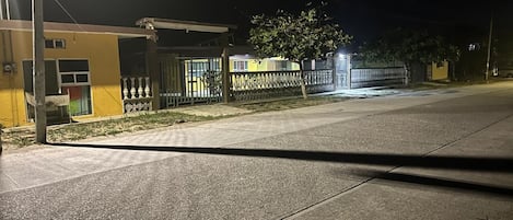 Una calle iluminada de noche, con un edificio que tiene una entrada protegida por una reja y un pequeño estacionamiento.