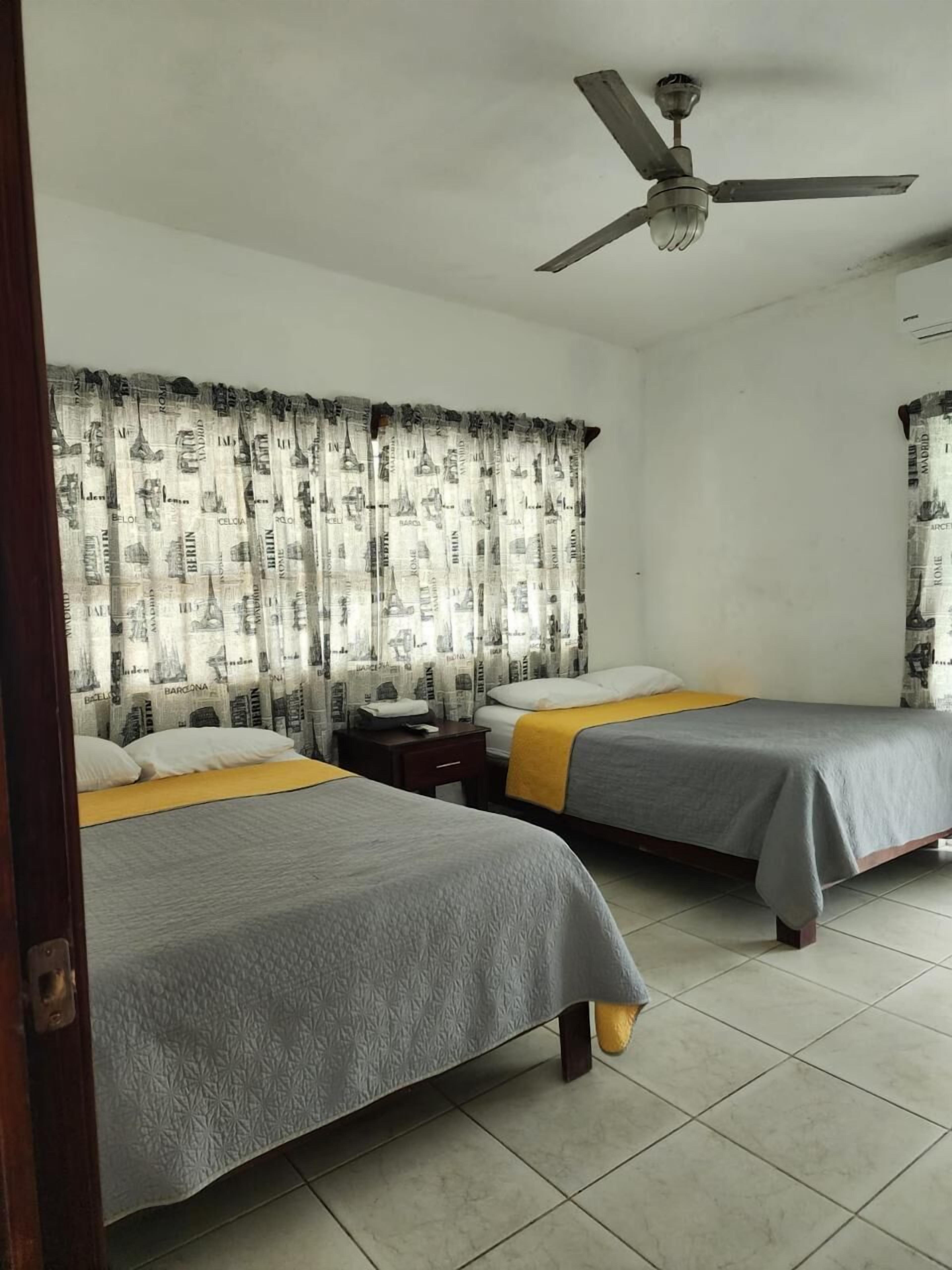Habitación de hotel con dos camas, ventilador de techo y cortinas con estampados de un paisaje urbano.