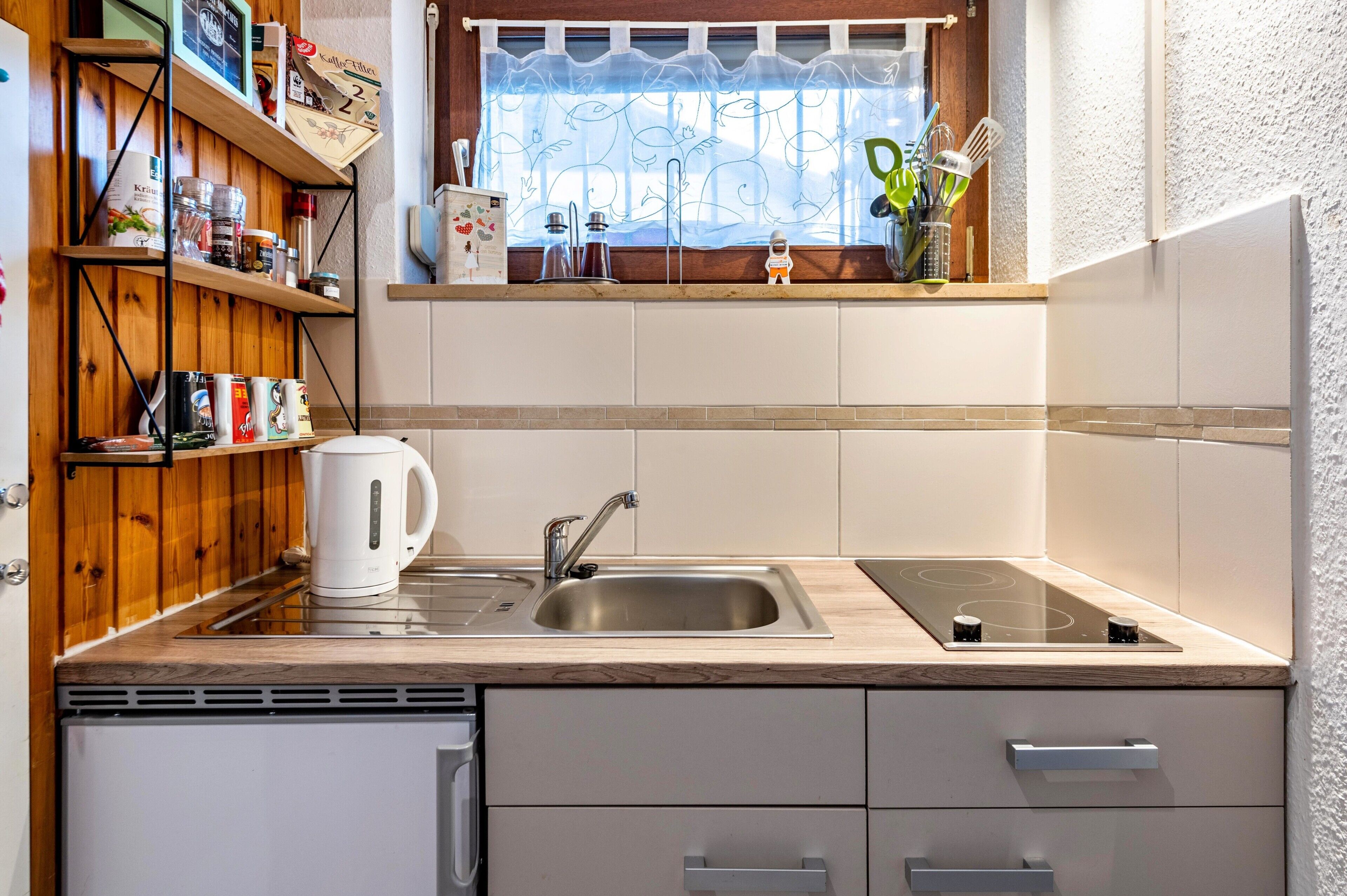 A kitchen with a sink, stove, wooden shelves, and a window with a patterned curtain.