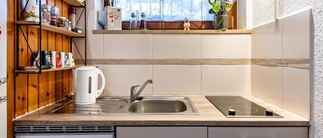 A kitchen with a sink, stove, wooden shelves, and a window with a patterned curtain.