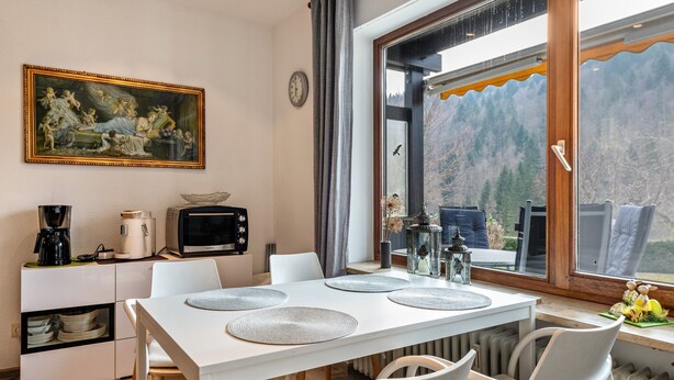 Zona pranzo con tavolo bianco, sedie, un quadro, un orologio e vista sulle montagne attraverso una finestra.