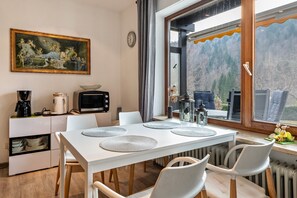 Comedor con una mesa blanca, sillas, un cuadro, un reloj y vistas a montañas a través de una ventana.