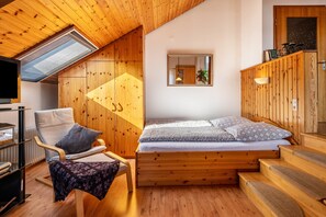 Une chambre confortable avec un plafond en pente, des murs en bois, un lit avec une literie à motifs, un fauteuil avec un coussin et une petite table recouverte d’une nappe à motifs.