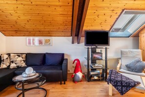 Un salon chaleureux avec un plafond en bois, un canapé noir, un gnome rouge, une télévision et un fauteuil.