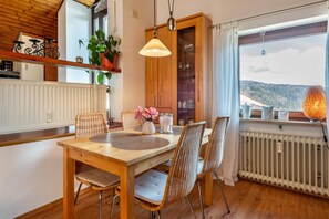 Un coin repas avec une table en bois, des chaises en osier, un luminaire suspendu et une vue sur un paysage montagneux par la fenêtre.