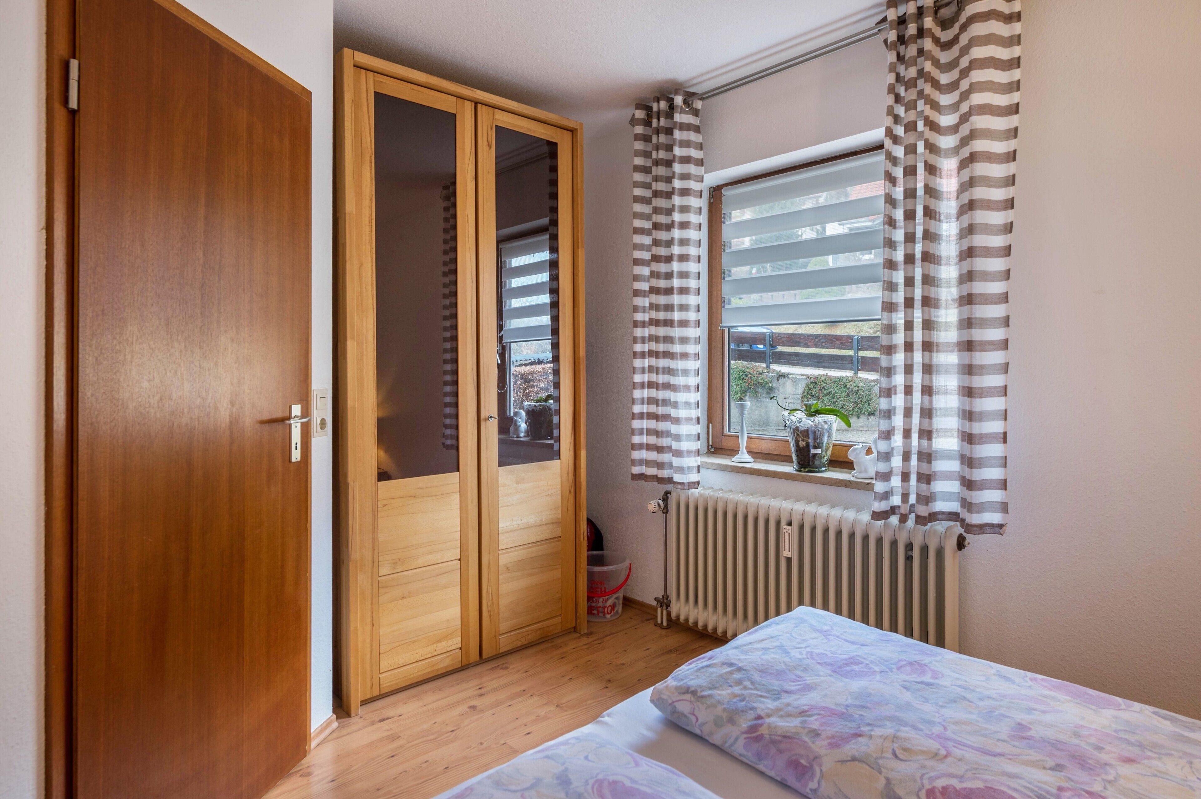 A bedroom with a wooden wardrobe, a window with striped curtains, a radiator, and a bed with floral bedding.