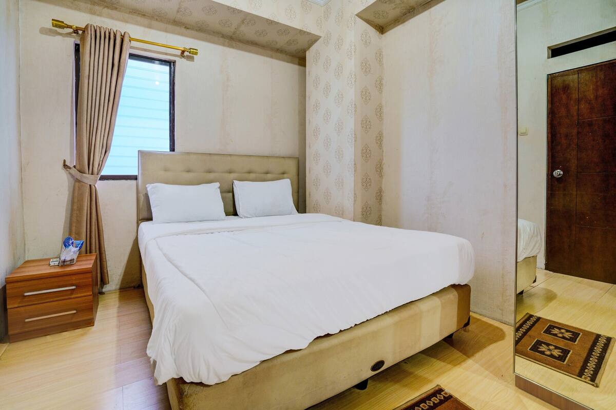 Un lit bien fait, avec une tête de lit, une table de chevet en bois dotée de tiroirs, une fenêtre avec des rideaux et une porte avec un miroir.