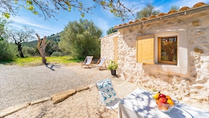 Un bâtiment rustique en pierre, doté d’une porte et d’une fenêtre en bois, avec un espace extérieur aménagé avec une table et des chaises, et un panier de fruits posé sur la table.