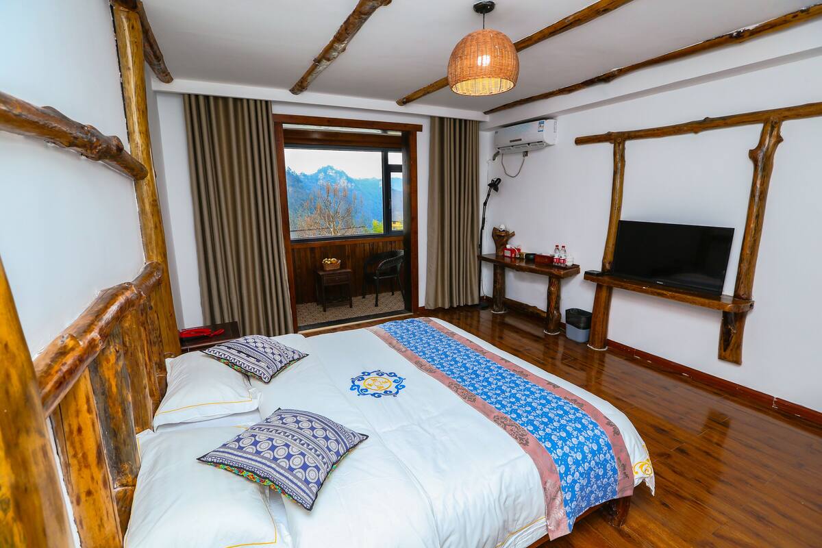 A bedroom with a wooden bed, a TV, a window with a mountain view, and a door leading to a balcony.