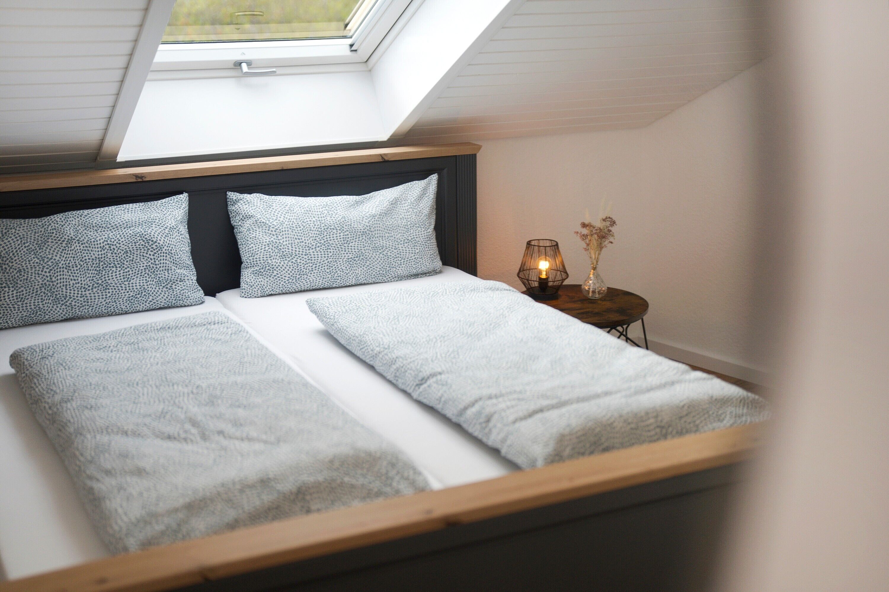 A bed with a skylight above, a bedside table with a lamp, and a vase with flowers.