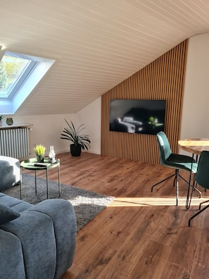 A modern living room with a sloped ceiling, wooden panel wall, a flat-screen TV, a dining table with chairs, a sofa, and a potted plant.