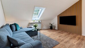 Ett modernt vardagsrum med ett sluttande tak, en stor platt-TV som är monterad på väggen, en grå hörnsoffa, ett litet bord med en vas och en krukväxt.