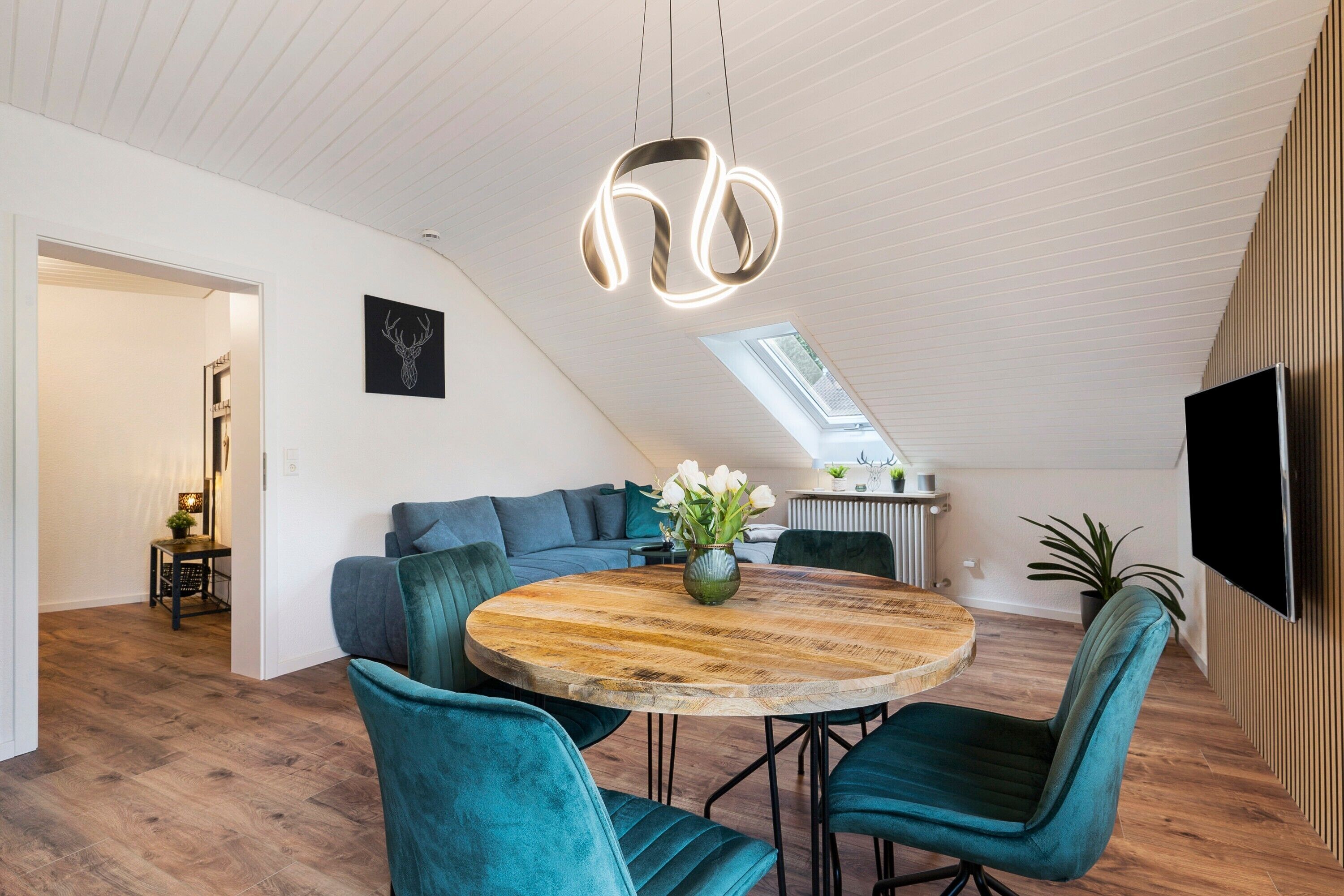 Un loft moderno con comedor que incluye una mesa redonda de madera y sillas color turquesa, una sala de estar con sofá y un tragaluz.