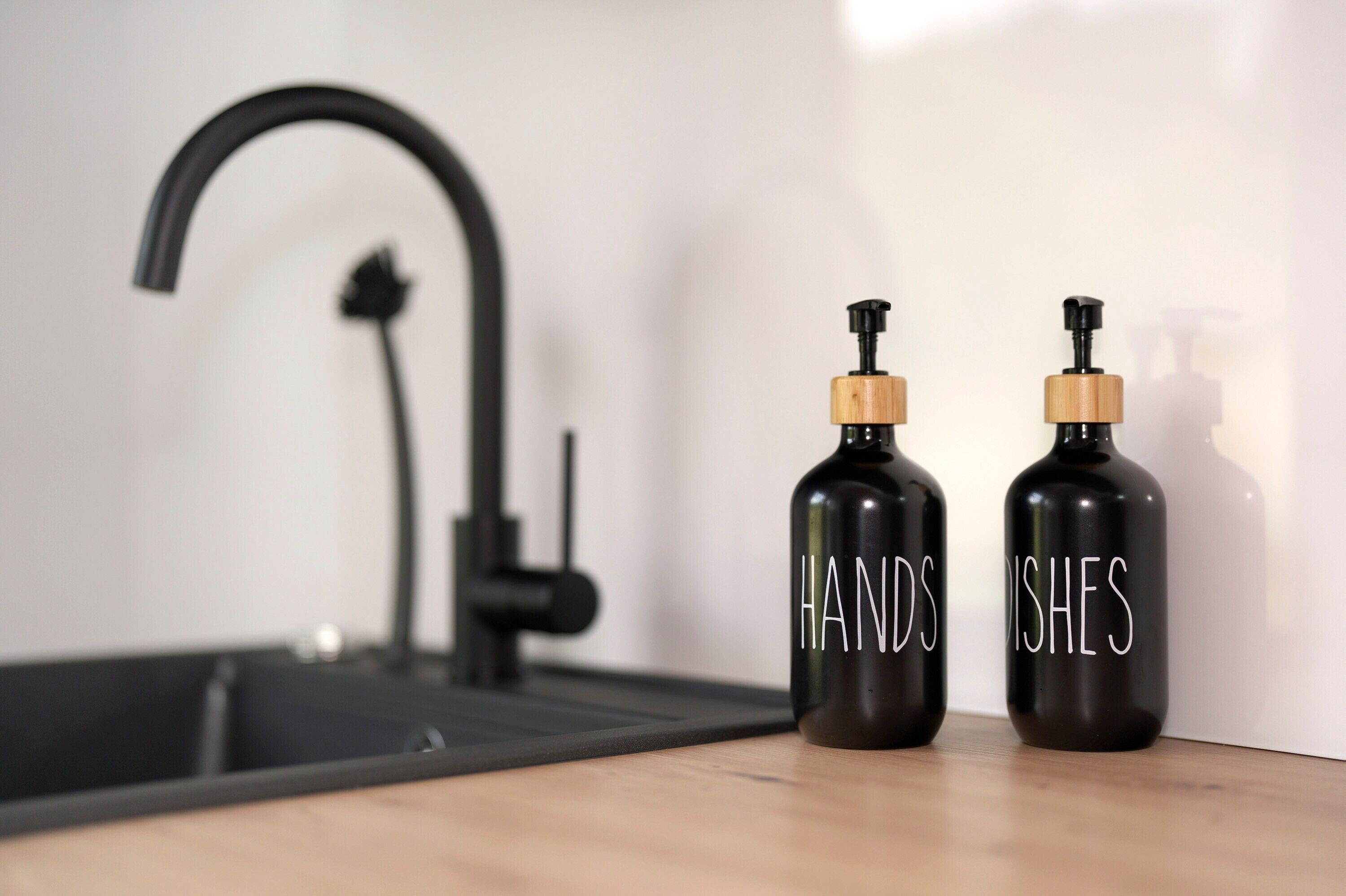 Twee zwarte flessen met houten dopjes, gelabeld 'HAND' en 'ISHES', op een houten oppervlak naast een zwarte kraan.
