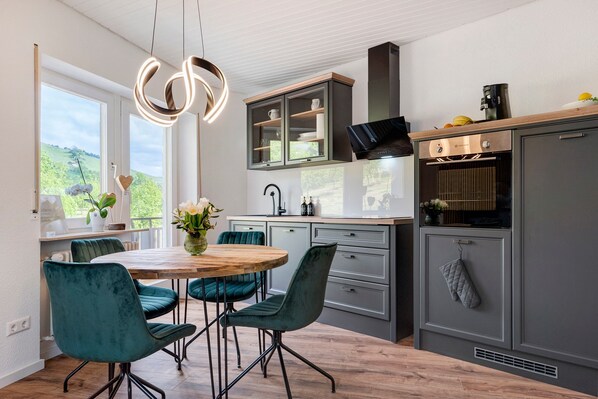 A modern kitchen with a wooden dining table, teal chairs, a built-in oven, and a view of greenery outside.