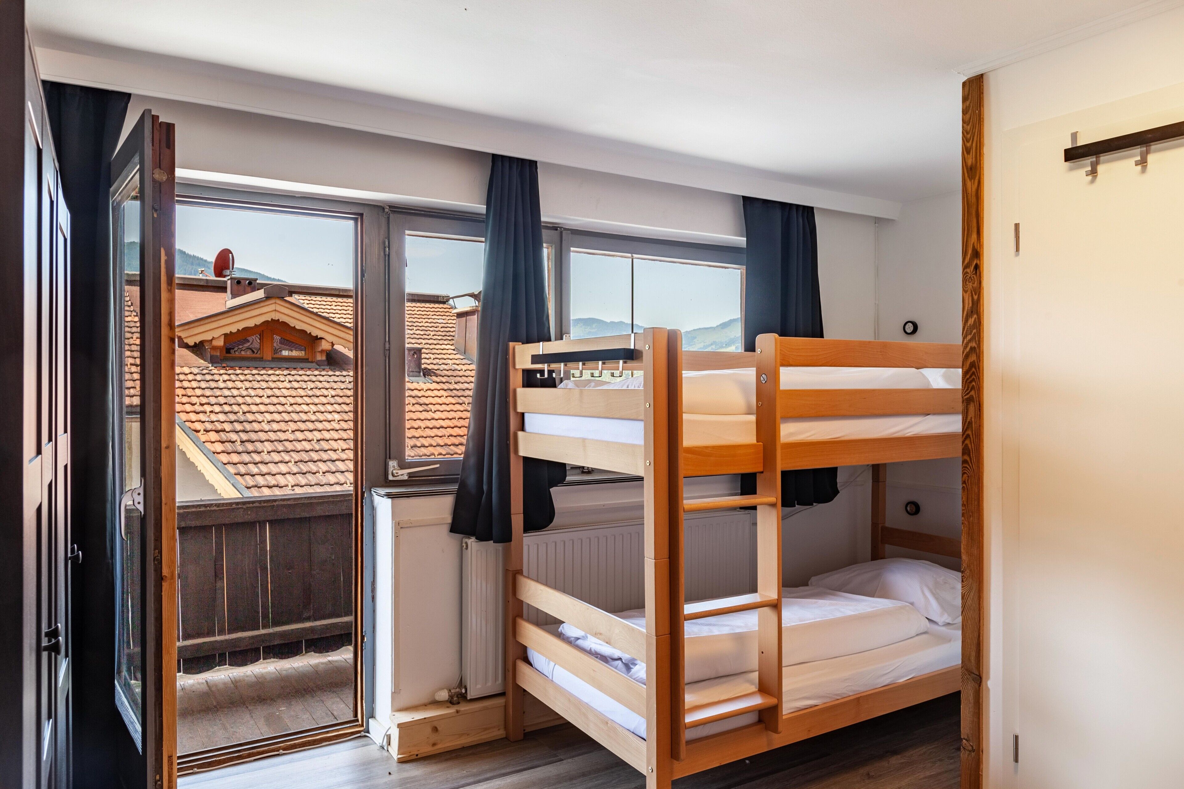 Een kamer met een stapelbed, een raam met gordijnen en uitzicht op een gebouw met een pannendak.