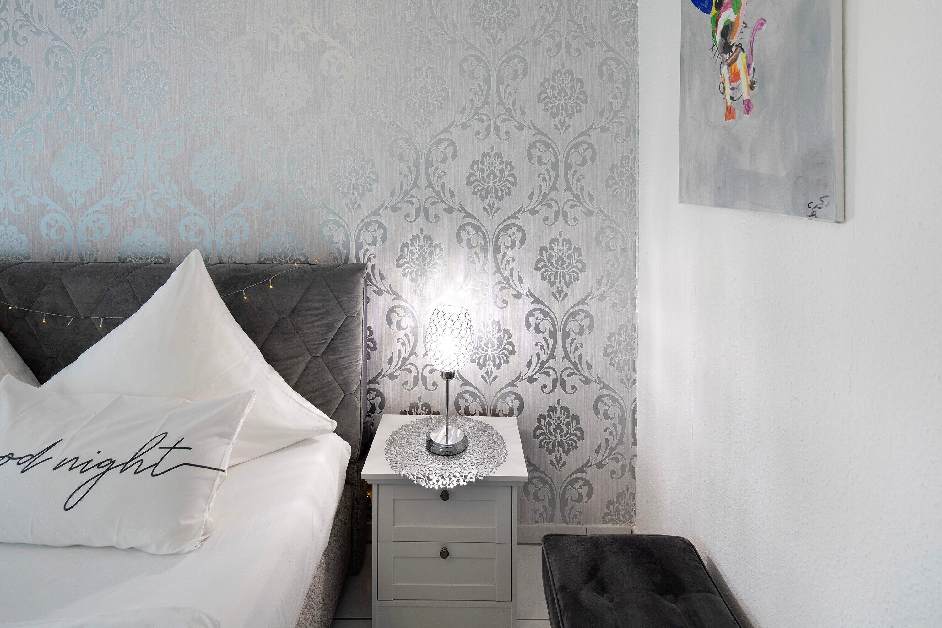 Un lit bien fait, avec un oreiller, une table de chevet avec une lampe et un mur orné d’un motif floral.
