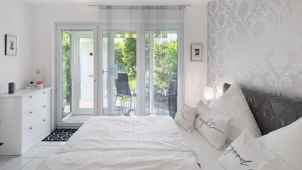Ein Schlafzimmer mit Bett, Kommode, Stuhl und Blick ins Freie.