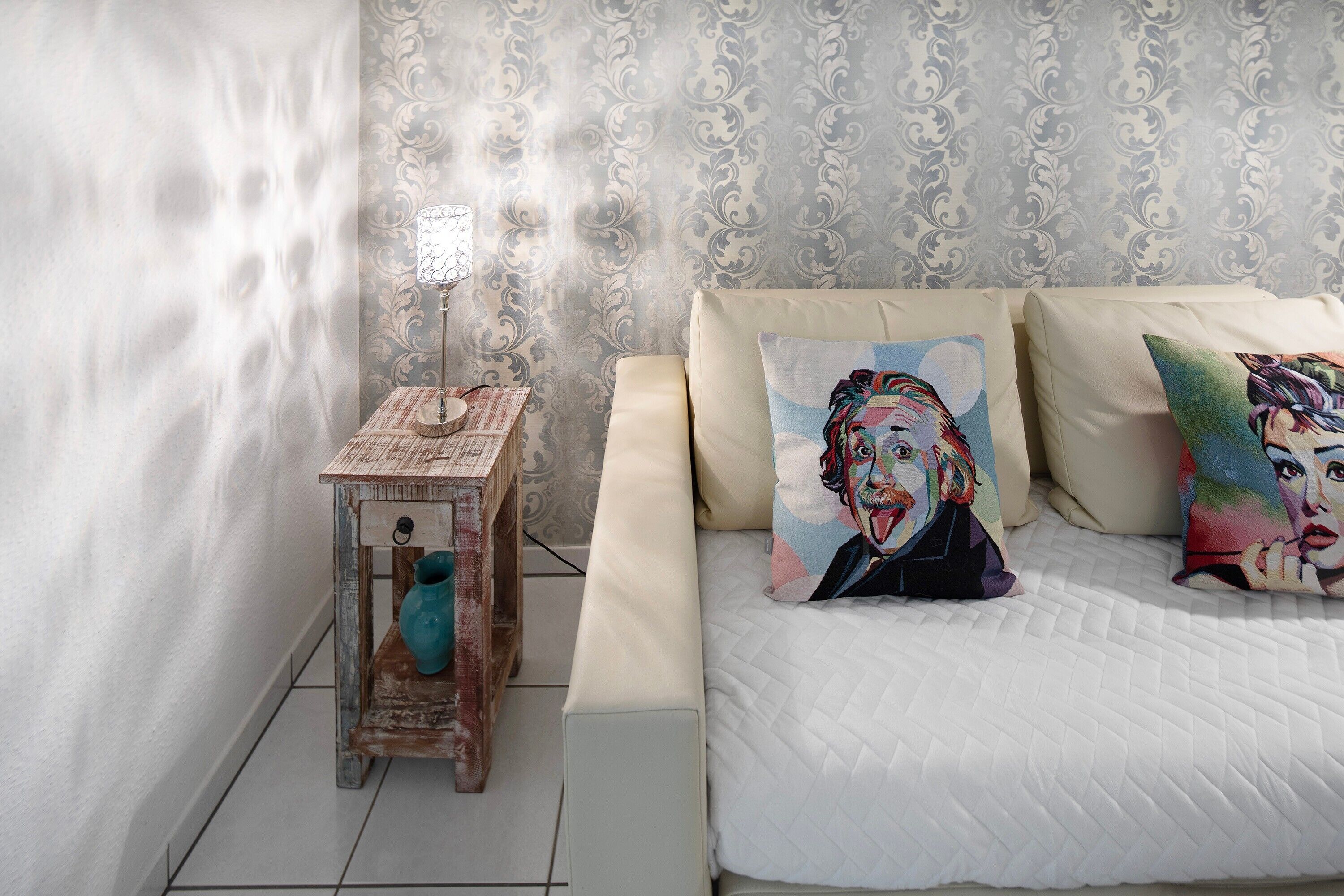 A bedroom with a bed, patterned wallpaper, a small wooden side table with a vase, and two pillows with pop art designs.