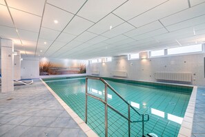 Piscina interior com chão em azulejo, escada de metal e vista para paisagem nevada através de uma janela.