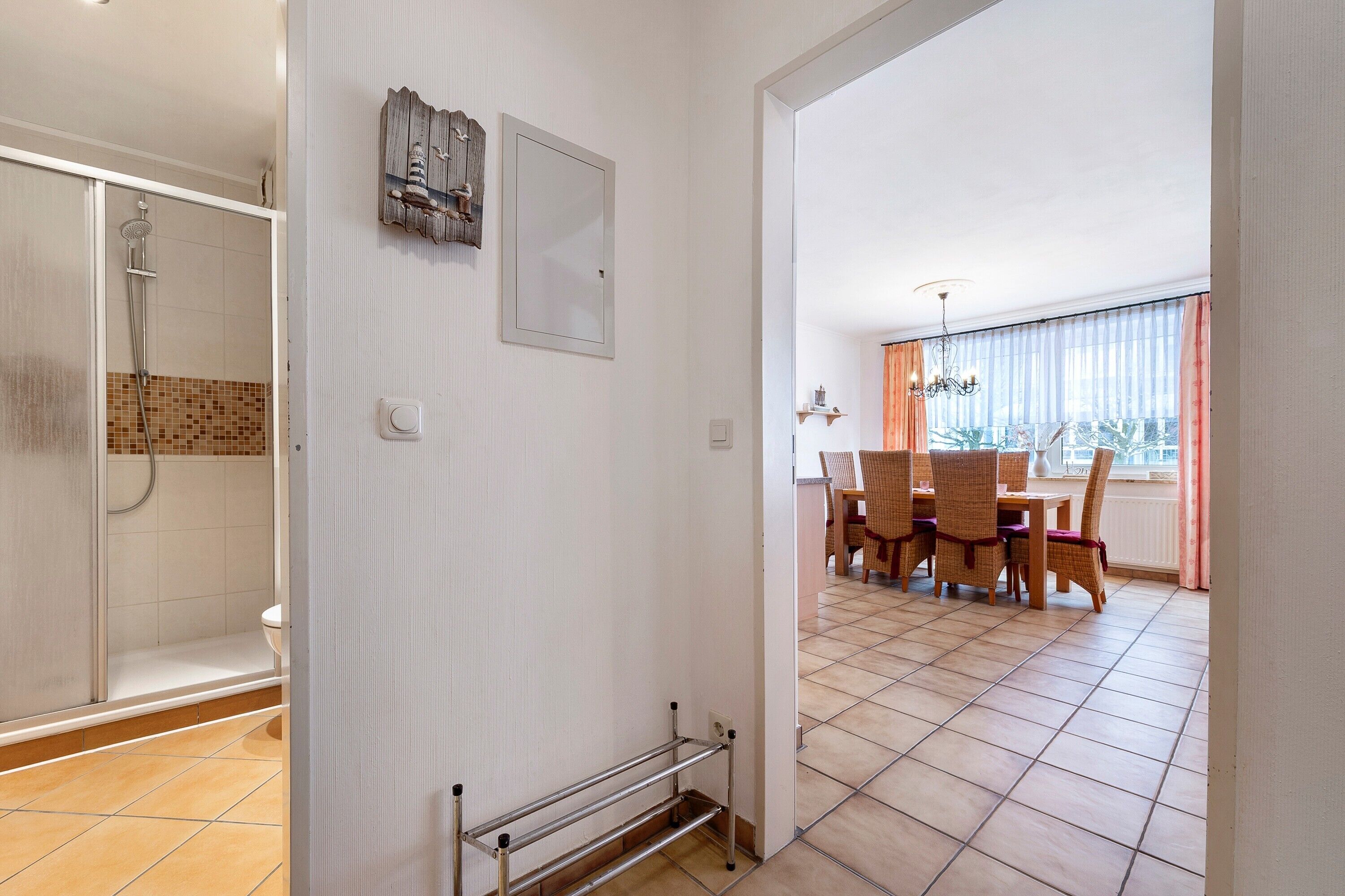 Ein Badezimmer mit Dusche, Toilette und Handtuchhalter. Durch eine offene Tür ist ein Essbereich mit Tisch und Stühlen erkennbar.