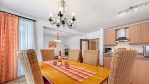 A dining area with a wooden table, wicker chairs, a chandelier, and a kitchen in the background.