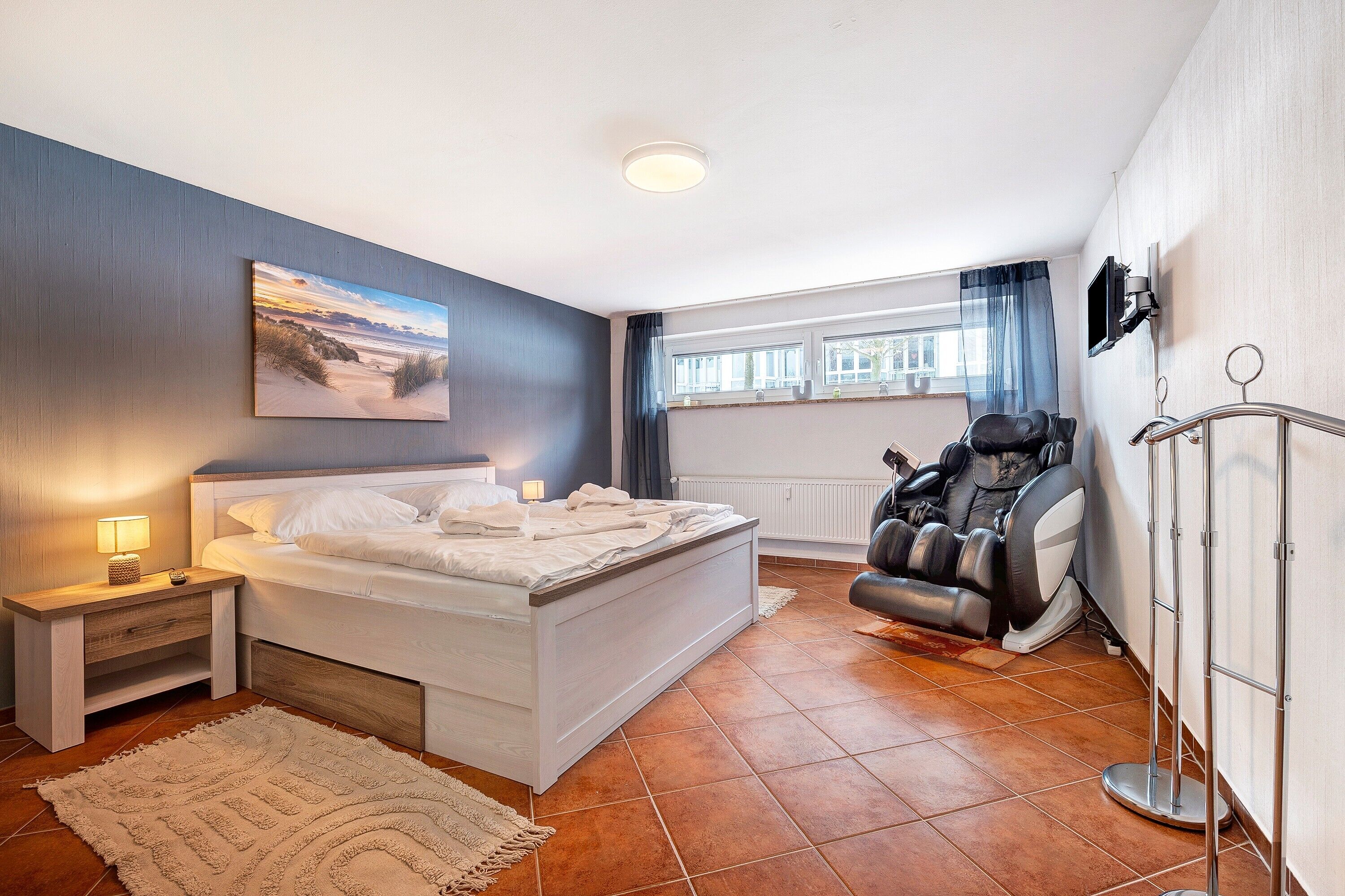 Ein Schlafzimmer mit Bett, Nachttisch, Lampe, Massagesessel und einem großen Fenster mit Vorhängen.