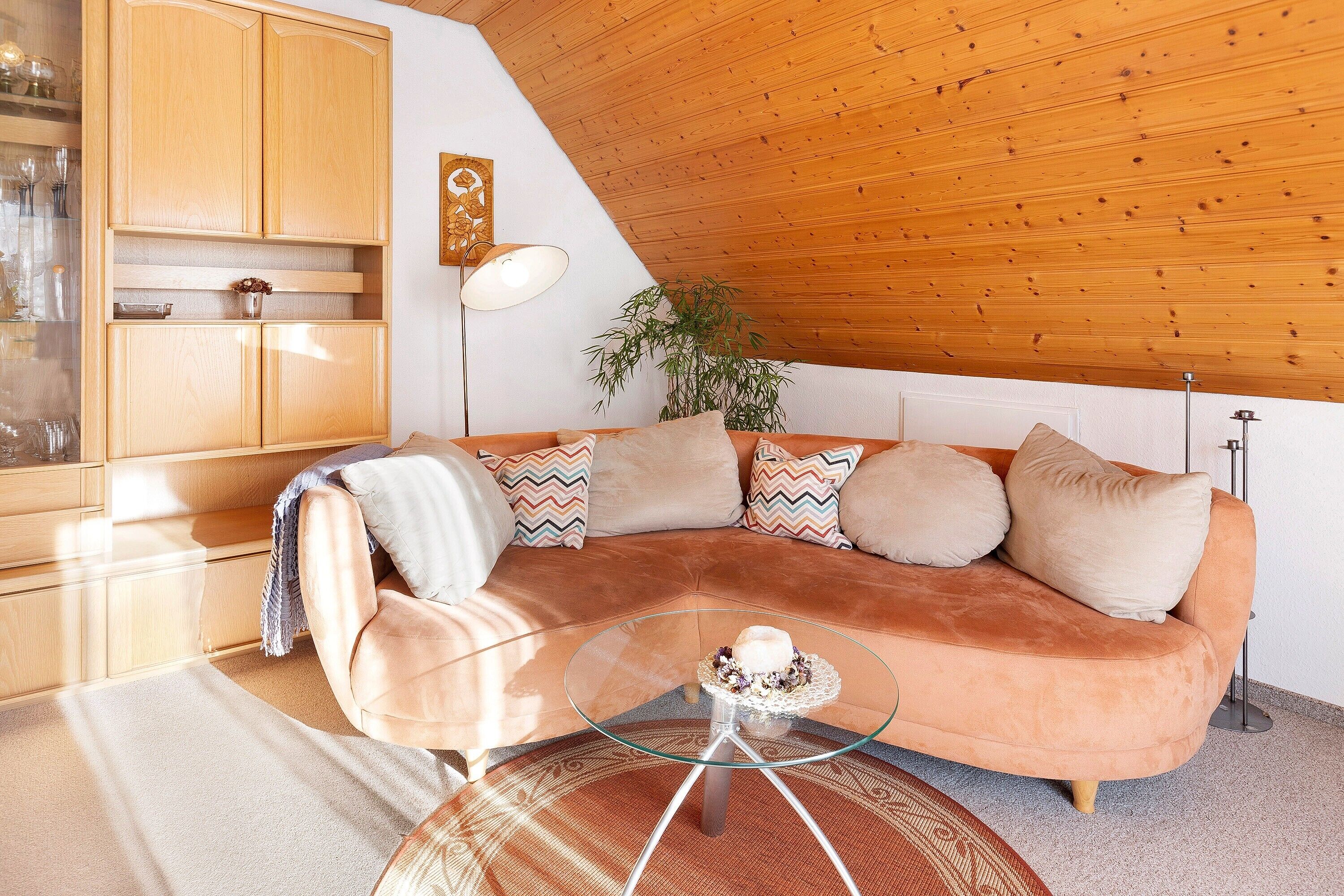 Een moderne woonkamer met een gebogen bank, een glazen salontafel, houten kasten en een plafond met zichtbare balken.