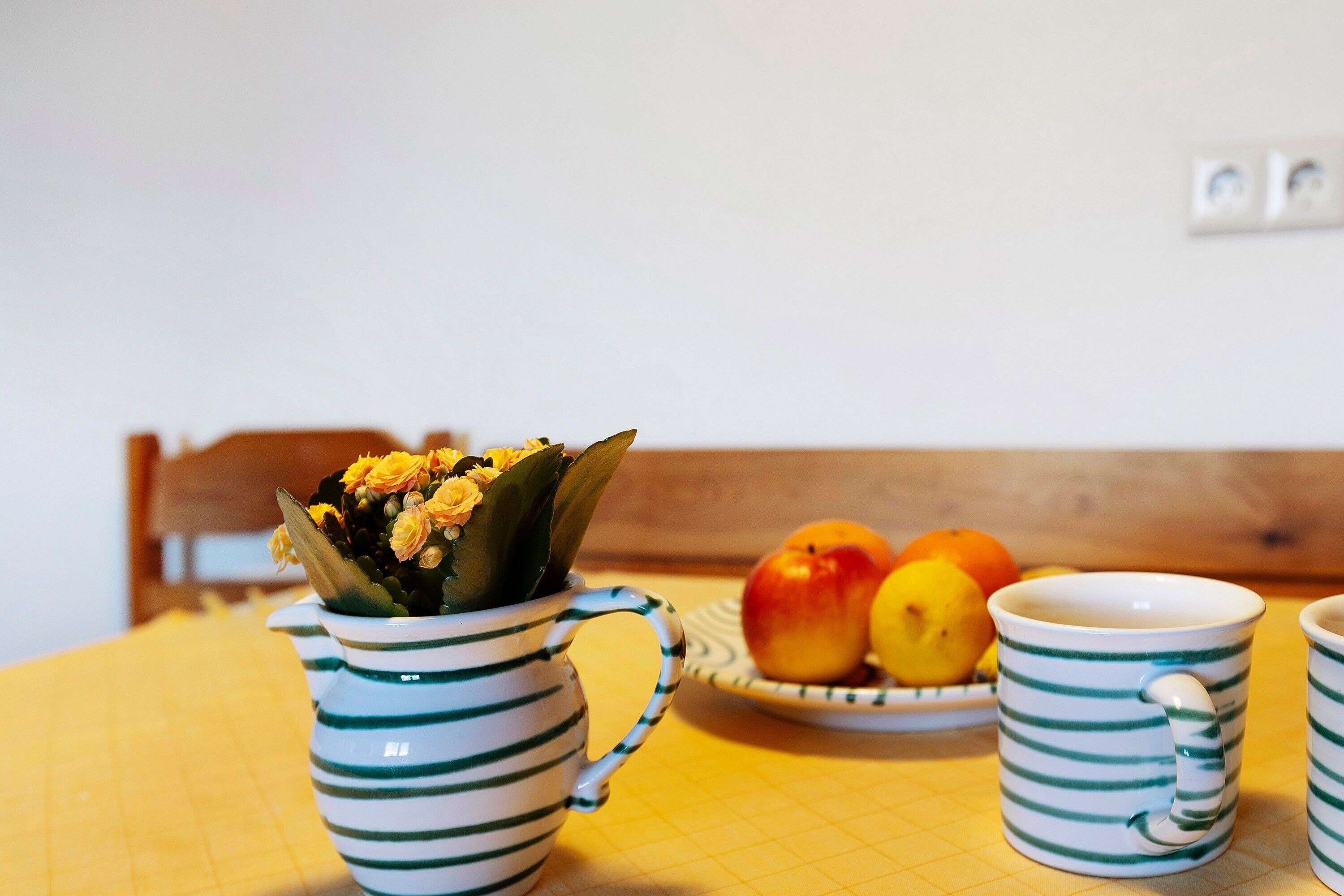 A table with a striped mug, a bowl of fruit, and a flower arrangement.
