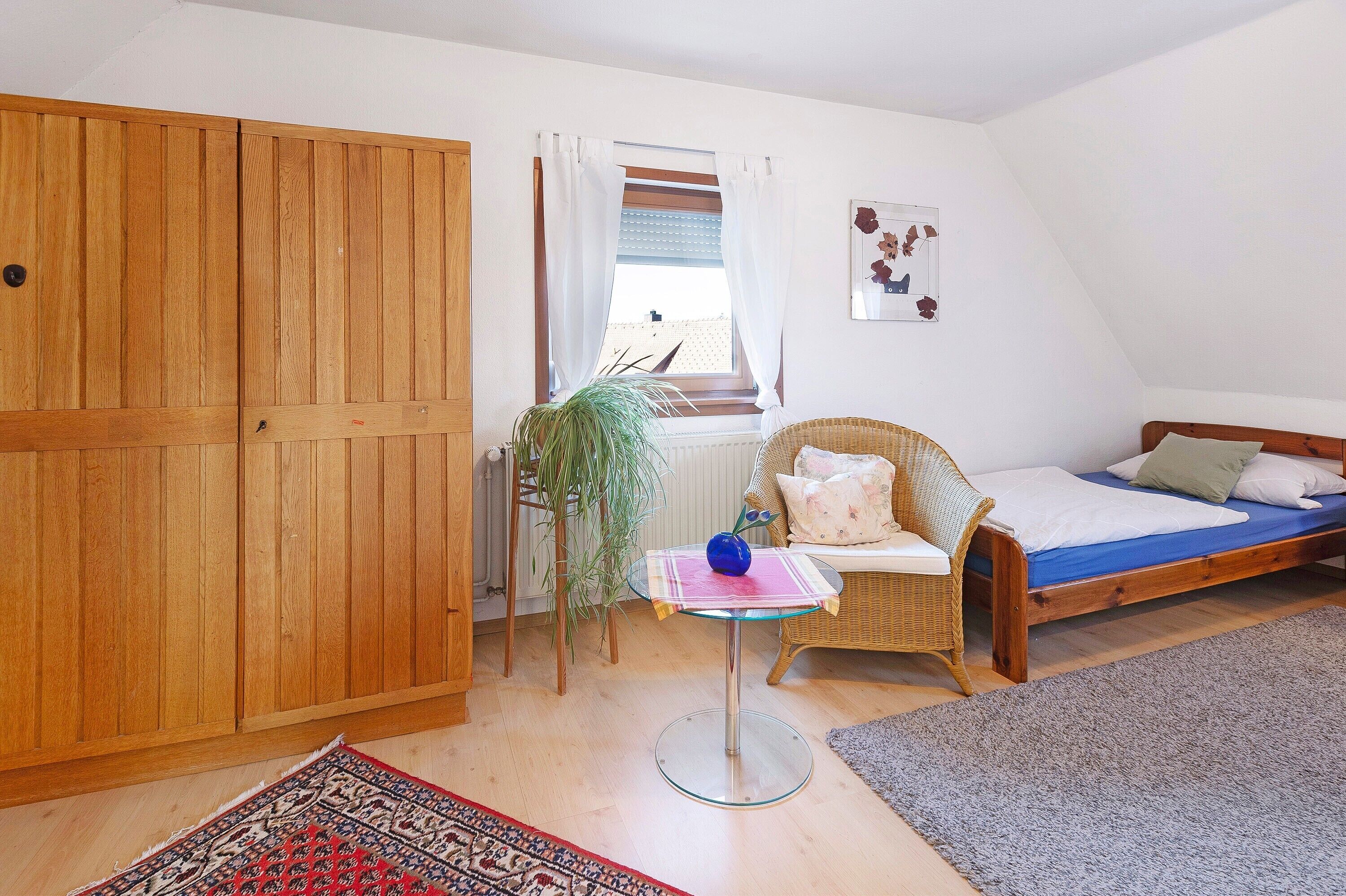 Ein Schlafzimmer mit einem Bett, einem Korbsessel, einem kleinen Tisch mit einer Vase, einem Holzschrank und einem Fenster mit Vorhängen.