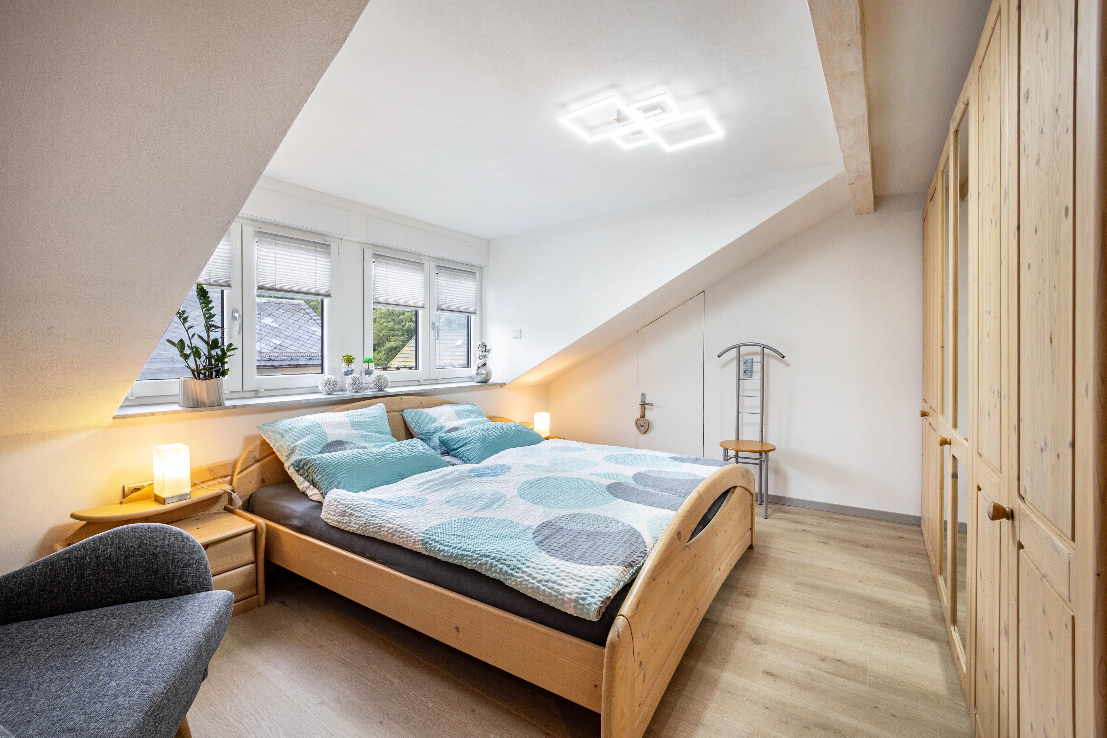 Een slaapkamer met een schuine kap, een bed met een gestreept beddegoed, een nachtkastje met een lamp, een stoel en een raam met jaloezieën.