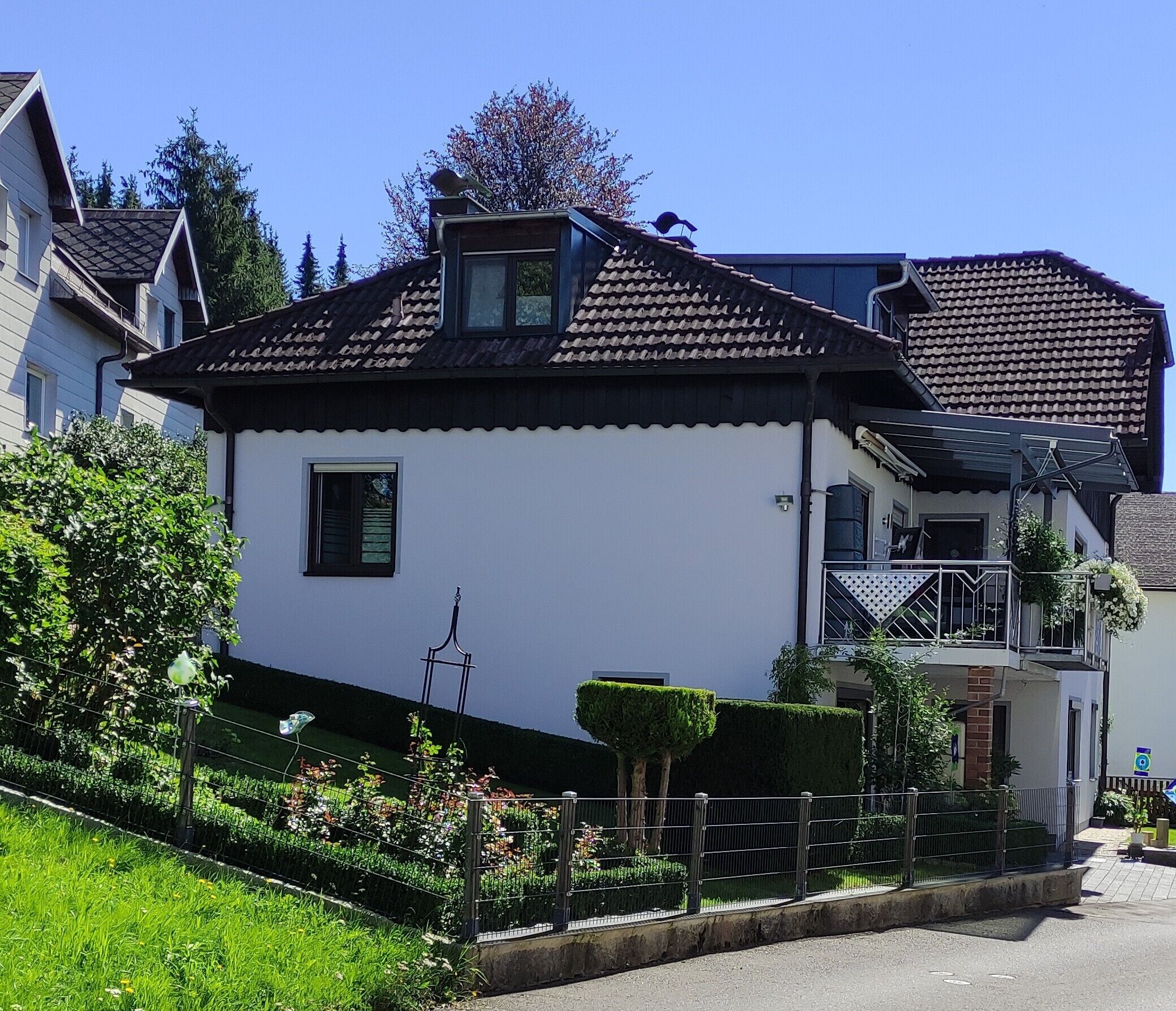 Une maison à deux étages, avec une façade blanche, un petit balcon et un jardin entouré d’une clôture.