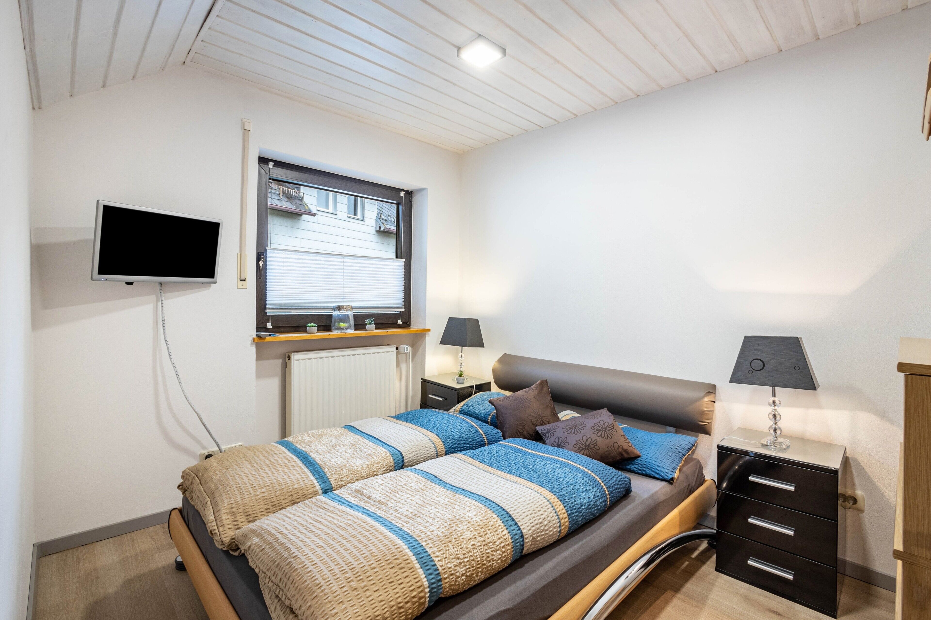 A bedroom with a bed, a TV mounted on the wall, a window, a bedside table with a lamp, and a dresser.