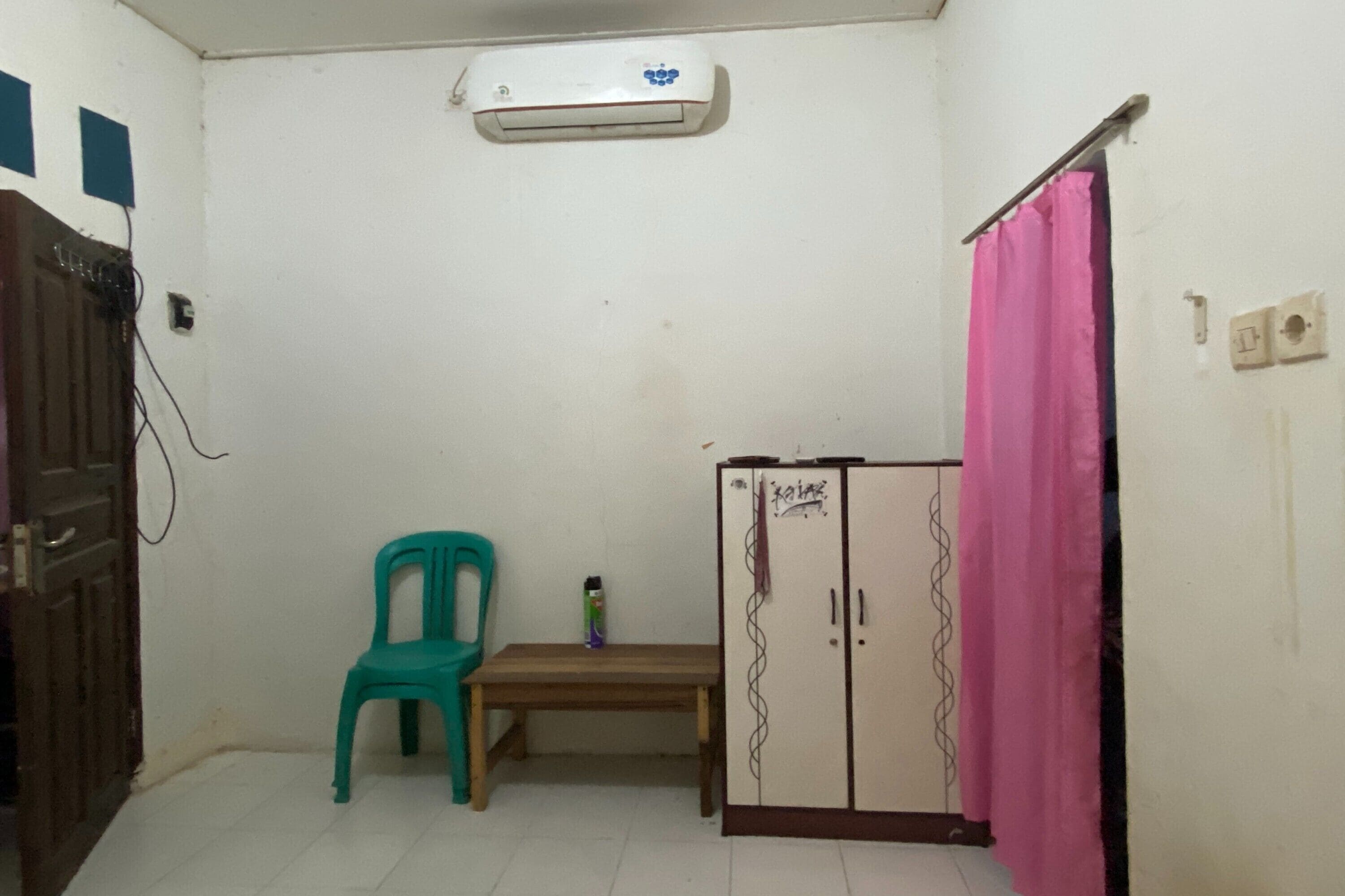 A small room with a wooden bench, a teal plastic chair, a white wardrobe with a pink curtain, and an air conditioning unit on the wall.