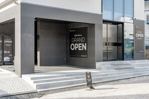A modern building with a large 'GRAND OPEN' sign, steps leading up to the entrance, and a 'NO PARKING' sign in front.