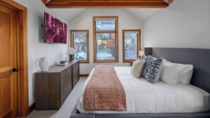 Un dormitorio con una cama grande, vigas de madera en el techo, un televisor en la pared y ventanas con vistas nevadas.